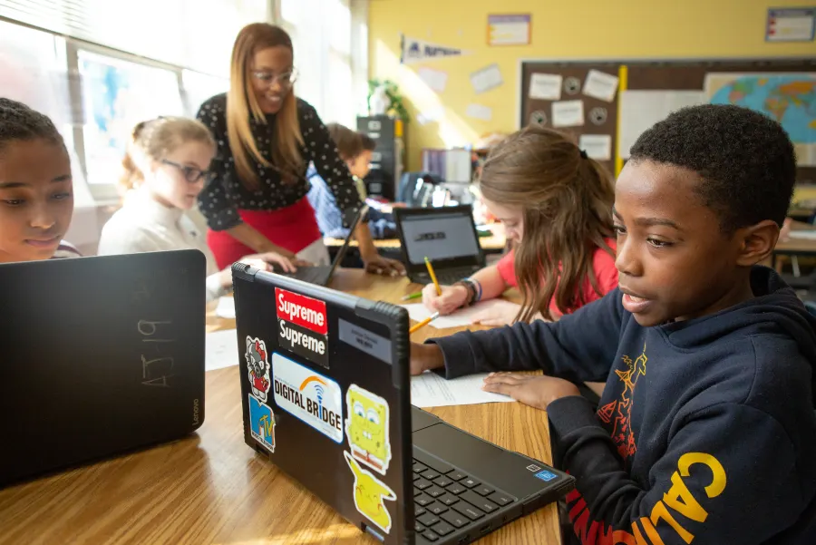 Middle school students research on laptops while their teacher helps